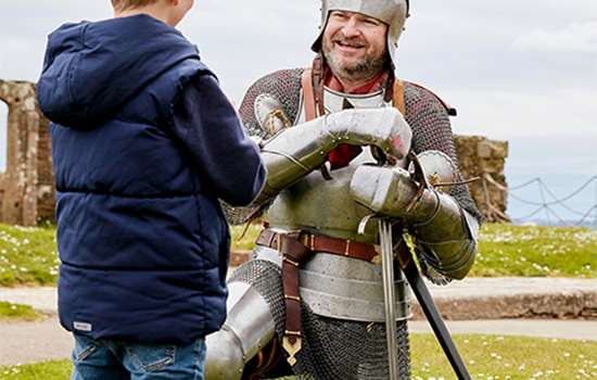 A man dressed as a knight bends on his knees to talk to a young visitor