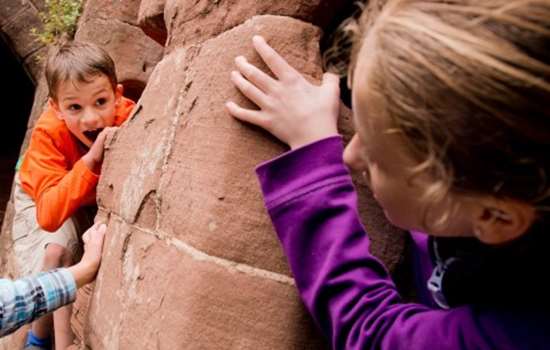 Children climbing and playing within castle walls. A boy in an orange sweatshirt peers behind a stone wall, while a girl in a purple sweatshirt looks at him, gripping the wall. 