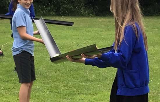 Two older children stand on a field, smiling, holding plastic guttering to pass a tennis ball between them. They both wear blue school uniform.