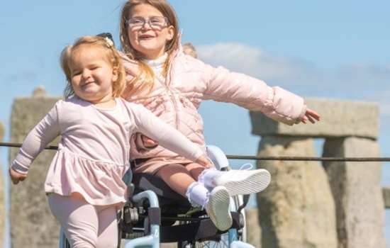 Two young girls are in front of Stonehenge, one is sitting in a wheelchair and the other stands beside her. They are both wearing pink and smiling. 