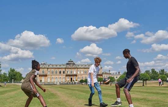 Three young men are kicking a football on grass in front of Wrest Park, a mansion in the style of a French chateau
