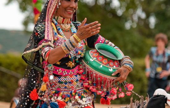 A woman in colourful traditional clothing dances outdoors, holding a decorated pot. She is smiling and wearing vibrant jewellery.
